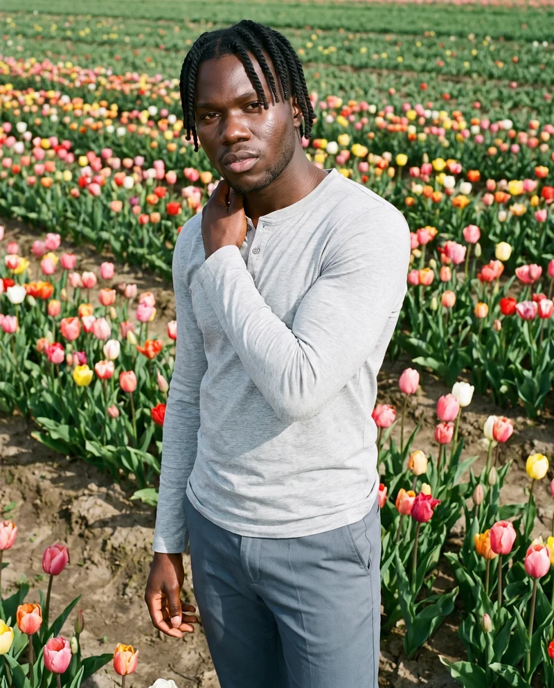 A 23-year-old African man is shown from the upper body at a front three-quarter angle, wearing a grey cotton Henley top. One hand rests on his neck, and he has a serious expression. A tulip field forms the backdrop.