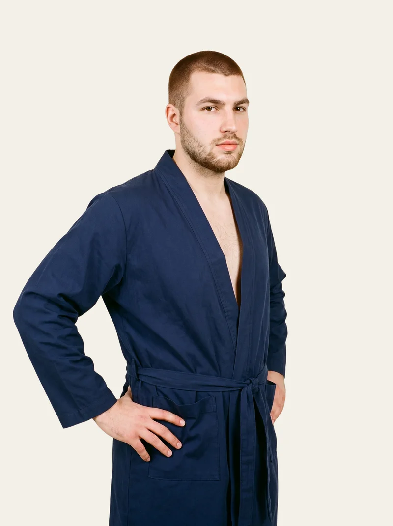 A 23-year-old Caucasian man wears a men's navy cotton dressing gown, shown in a three-quarter-length frame from a front three-quarter angle with his hands on his waist and a soft facial expression. The alabaster flat-color backdrop keeps the focus on the garment.