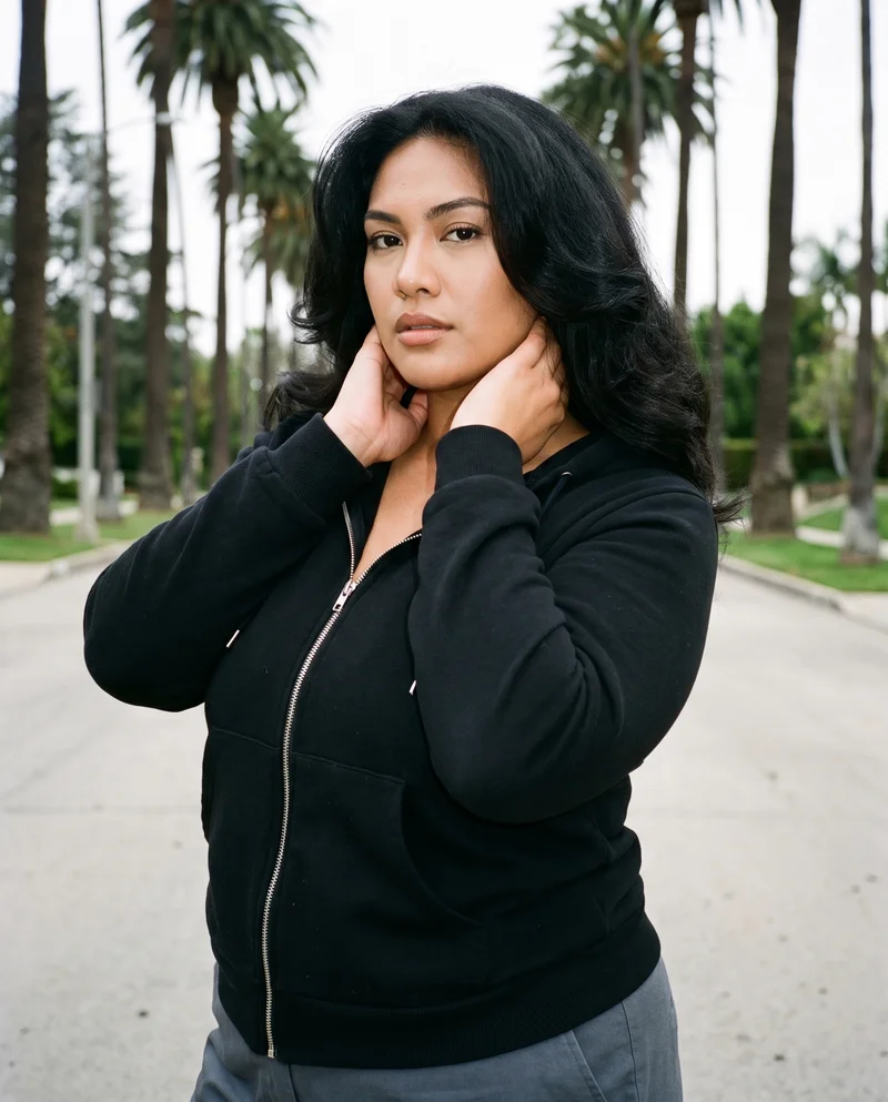 A 22-year-old Southeast Asian woman wears a black zip-up hoodie in an upper-body front three-quarter view, with her hands on her neck and a natural expression. A Palm Mansion Boulevard backdrop is visible behind her.