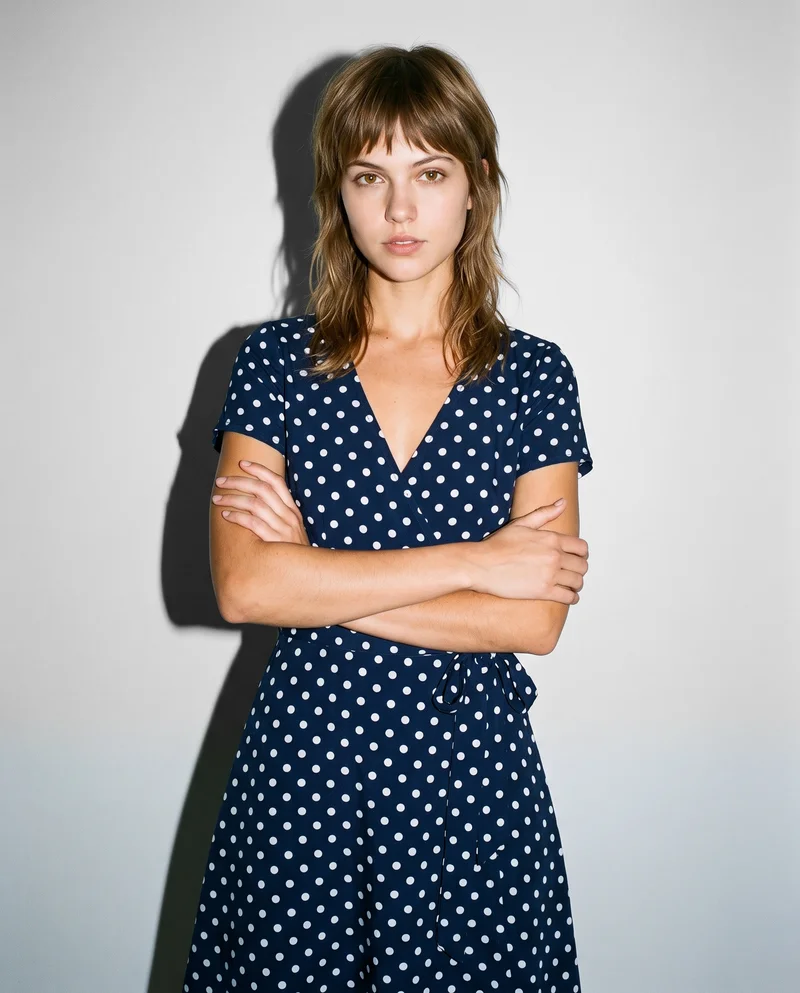 A 23-year-old Caucasian woman wears a polka-dot wrap dress in a front-facing, three-quarter-length view with her arms crossed and a confident expression. A fog grey studio-wall backdrop fills the background.