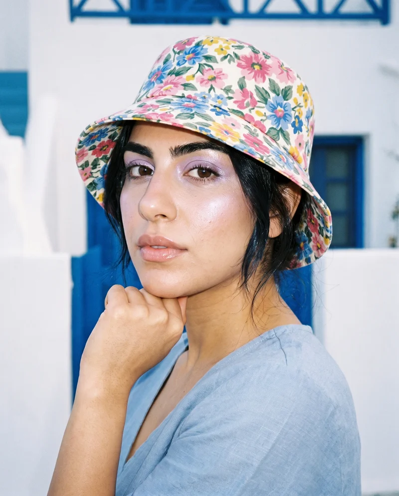 A 23-year-old Arab woman wears a women's floral print bucket hat in a bust composition, shown from a front three-quarter angle with her chin resting on her knuckles and a neutral expression. A Greek Villa White Blue backdrop is visible behind her.