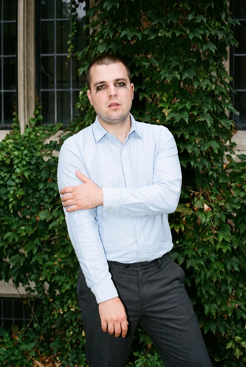 A 34-year-old Caucasian man wears a light blue Oxford shirt in a front-view, three-quarter-length composition. He is shown holding his arm with a soft facial expression, against an ivy-covered building backdrop.