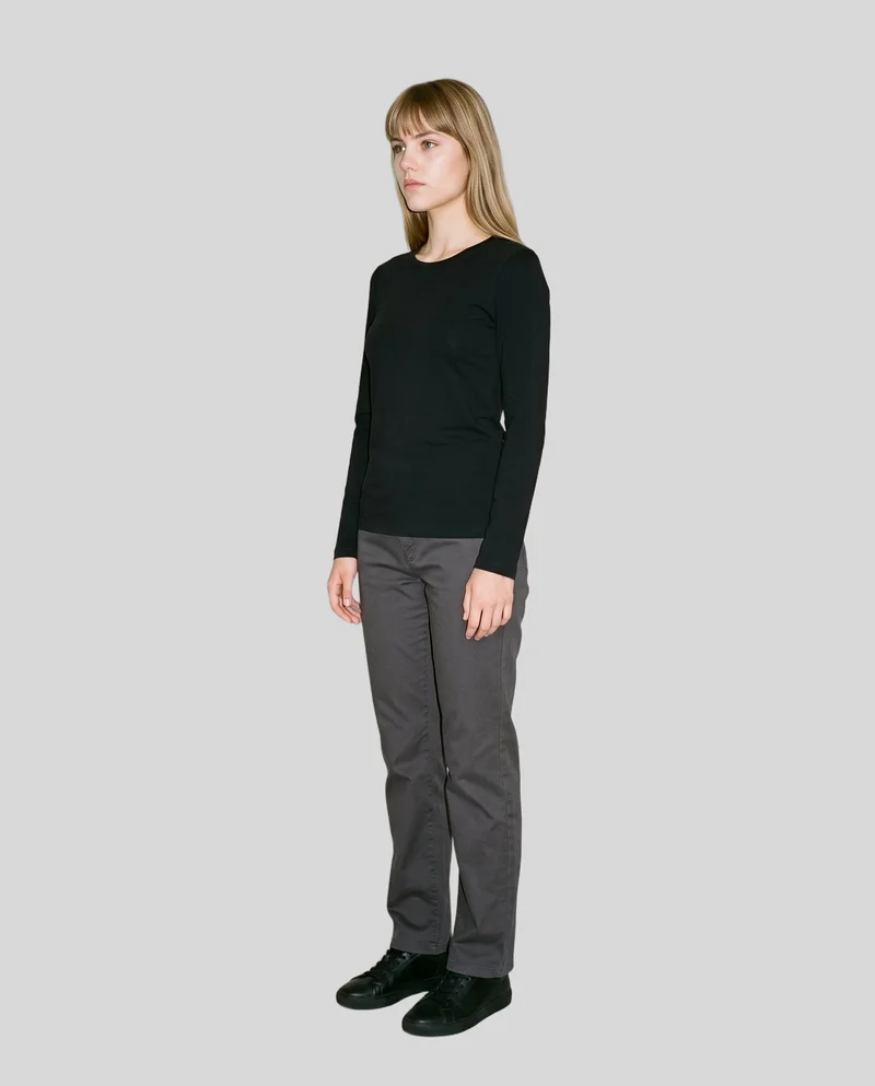 An 18-year-old Caucasian woman stands straight in a full-body, angled front view wearing a women's black long-sleeve tee. Her expression is neutral, with a pearl grey flat-color backdrop.