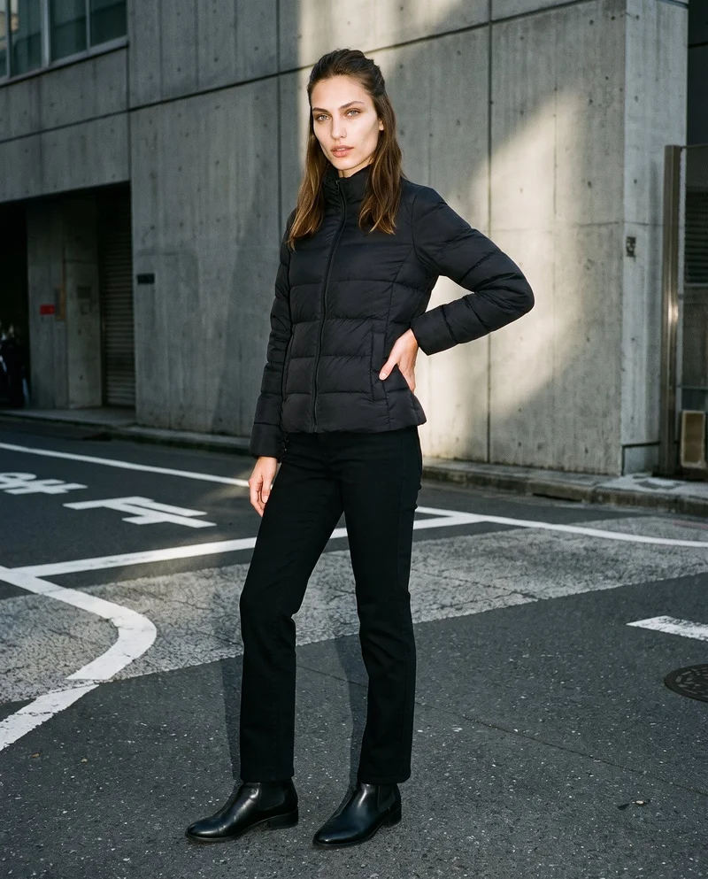 A 19-year-old Turkish female model wears a women's black puffer jacket in a full-body, angled-front view with one hand on her hip and a sultry expression. A Tokyo Crossing backdrop is visible behind her.