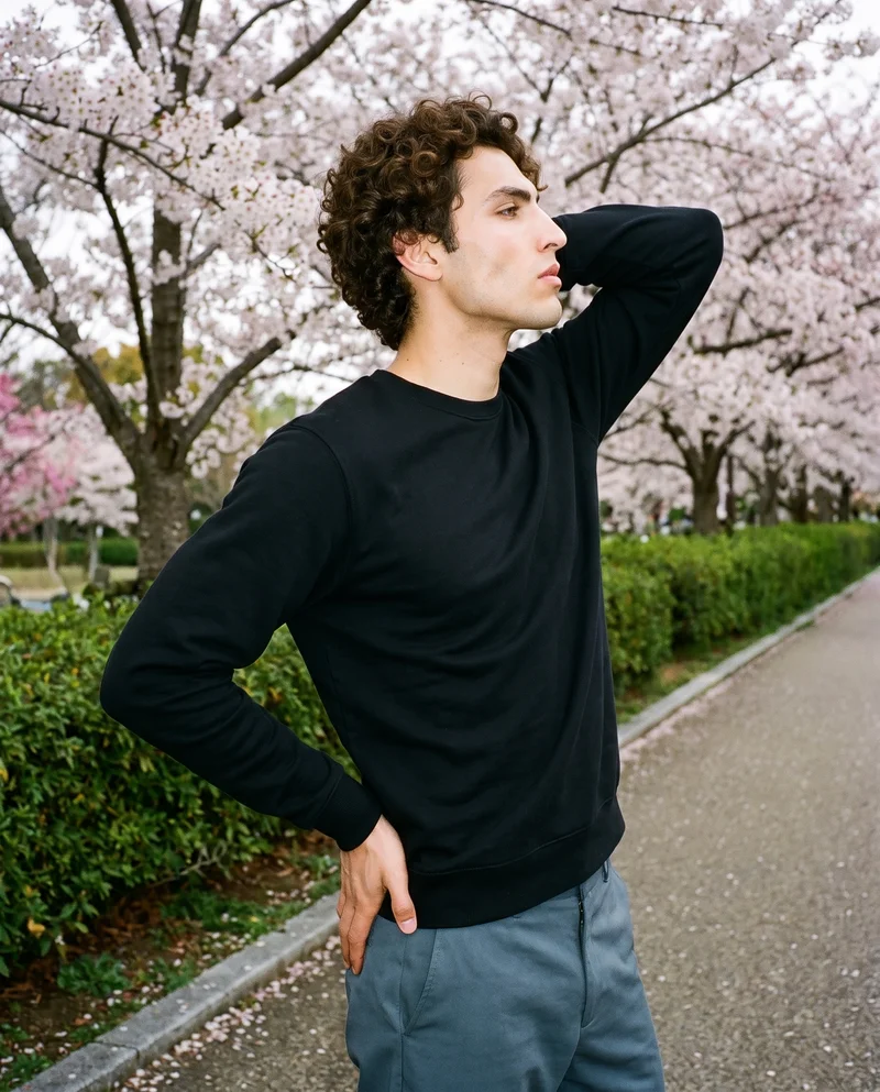 A 25-year-old Hispanic man is shown from the upper body in side view wearing a black crewneck sweatshirt. His hand is positioned behind his neck, and he has a fierce facial expression. A Cherry Blossom Park backdrop is visible.