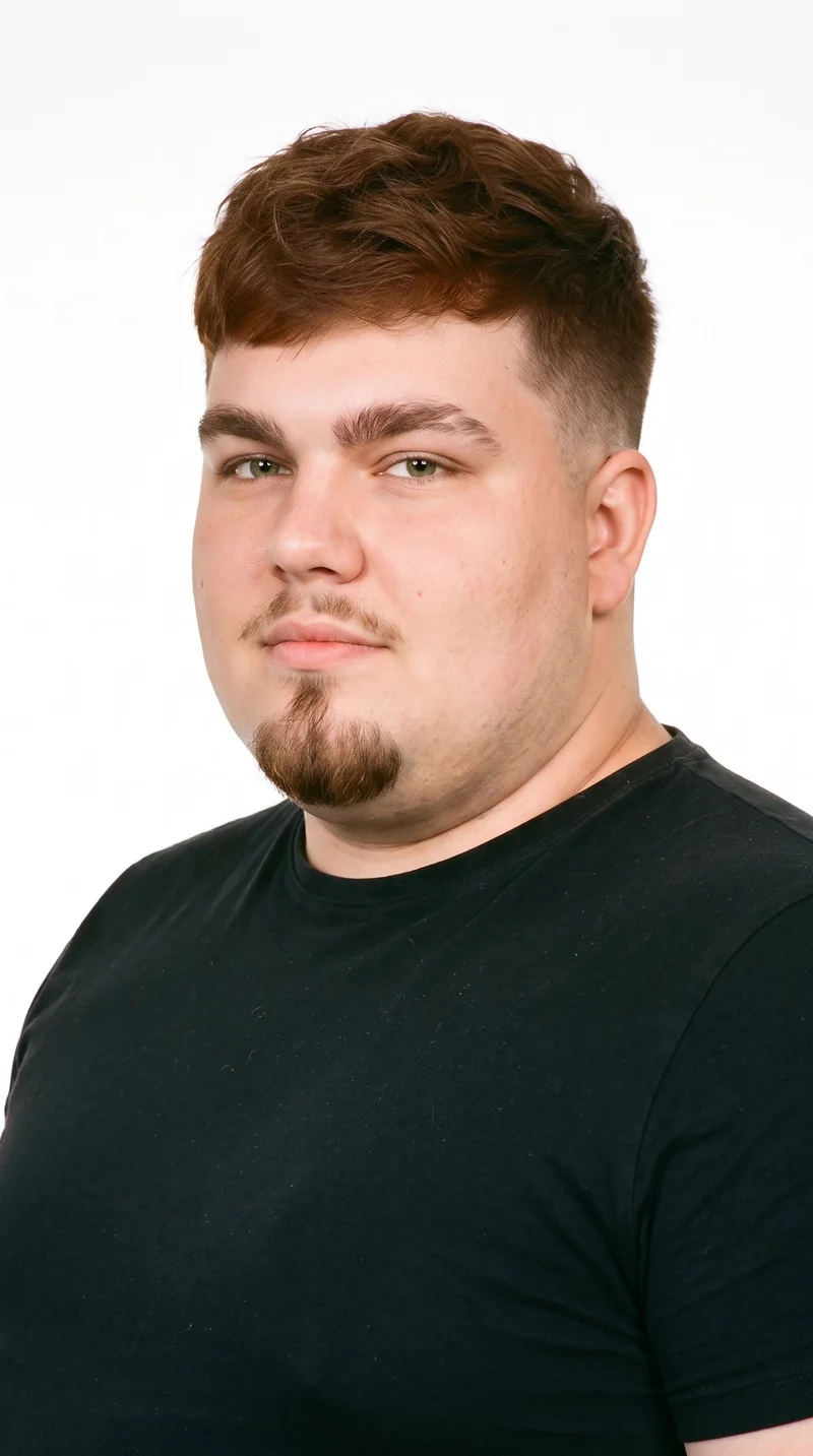 This 21-year-old Caucasian man has lightly tanned fair skin with a peach undertone. His face has smooth, even, softer features in a refined, balanced shape. He has thick eyebrows, green eyes, and full lips. His chestnut-brown hair has a reddish undertone and is styled in a short textured crop with layered texture on top and closely cut sides. A compact goatee is centered on his chin, while his cheeks are clear.