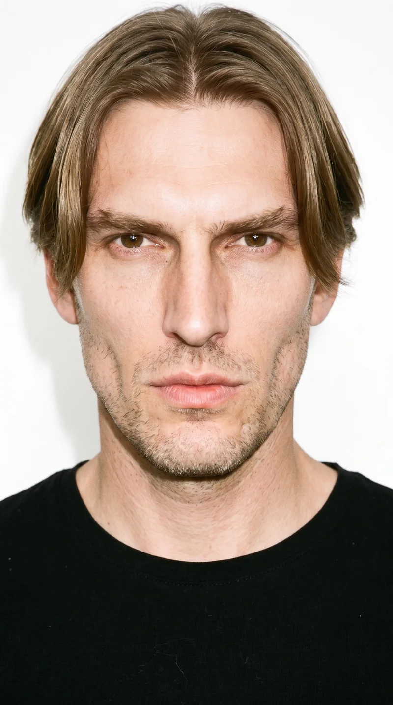 A 41-year-old Caucasian man with fair skin and a peach undertone has a gaunt face with lean features, hollowed cheeks, and sharply sculpted bone structure. His thick eyebrows frame brown eyes, and he has full lips. His dark-blond hair is divided at the centre and falls evenly to both sides, while fine, closely trimmed stubble lightly covers the beard area.