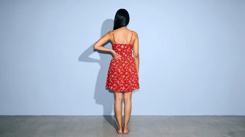 A 26-year-old Indian woman wears a red floral sundress, shown from the back in an environmental frame with one hand on her lower back. A powder blue studio-wall backdrop fills the setting.