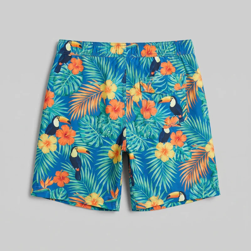 Tropical print board shorts laid flat with the back side facing up. The men's swim shorts have a mid-rise elastic waistband, straight knee-length legs, a rear patch pocket with a flap on the wearer's right side, and an all-over print of palm leaves, hibiscus-style flowers, and toucans.