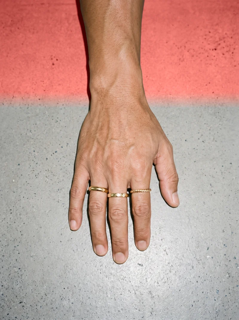 A hand and wrist are shown from above with fingers draped while wearing a men's gold stacking ring set. The coral pink studio-wall backdrop fills the background.