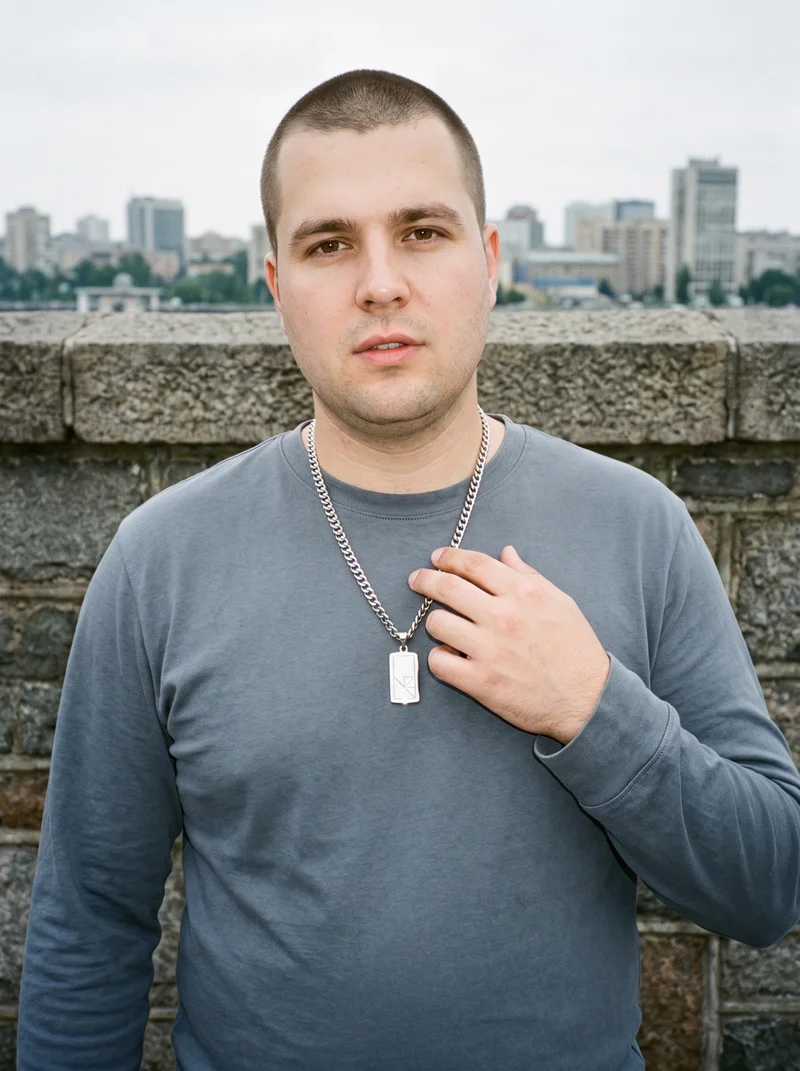 A 34-year-old Caucasian man is shown from the front in an upper-body view, with his fingers at his chest and a soft facial expression. He is wearing a men's silver pendant necklace, with an island causeway city skyline backdrop behind him.