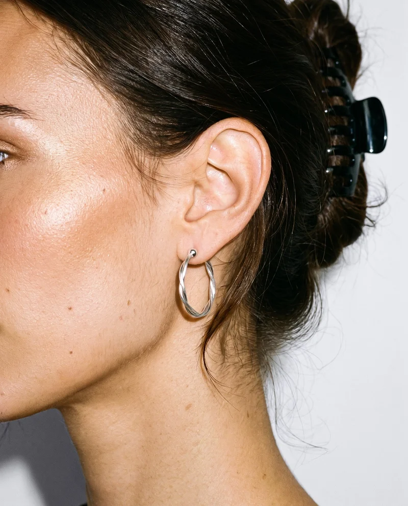 A silver ear cuff is shown on a woman's ear in side view, with her head held level. The ear is framed against a porcelain studio backdrop.