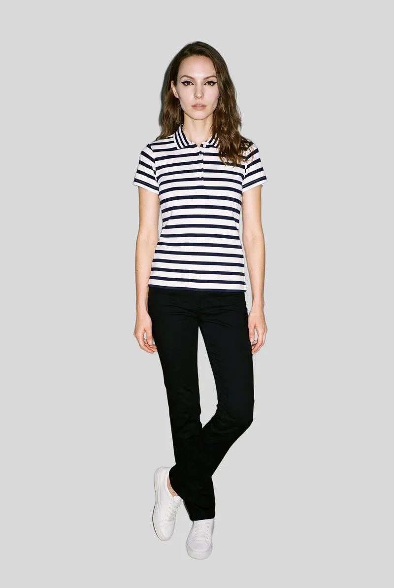A 26-year-old Caucasian woman is shown full-length from the front wearing a women's striped cotton polo shirt. She stands with her ankles crossed against a pearl grey backdrop, with a confident expression.