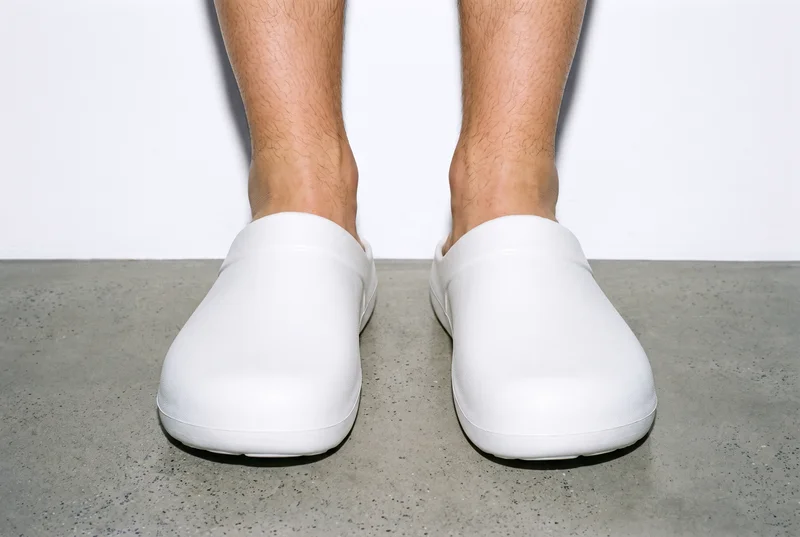 A pair of men's white clogs is shown from the front with both feet together. The shoes are set against a porcelain studio-wall backdrop.