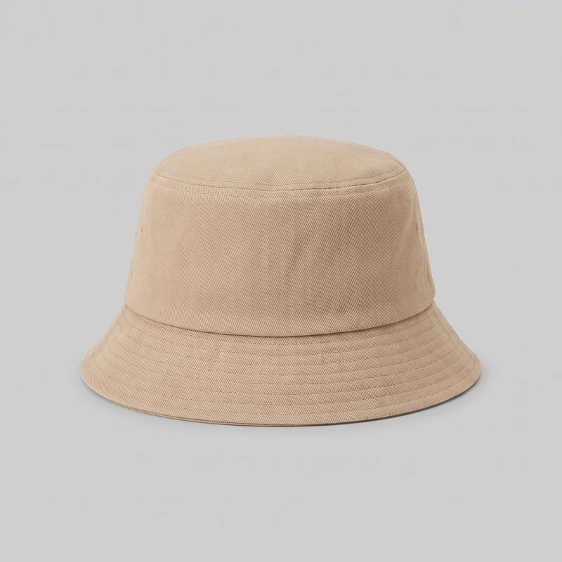 A standalone beige bucket hat is shown from a slightly elevated front angle. Its flat circular crown, straight cylindrical side wall, downward-sloping brim, crown seam, subtle panel seams, and small side ventilation eyelets are visible, along with multiple concentric rows of stitching across the brim and a finished outer edge.