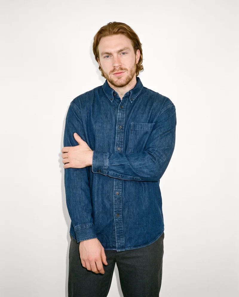 A 25-year-old Caucasian man wears an oversized denim shirt in a front-facing, three-quarter-length view. He holds one arm in a natural pose and has a natural facial expression, with a Chantilly Lace studio-wall backdrop.