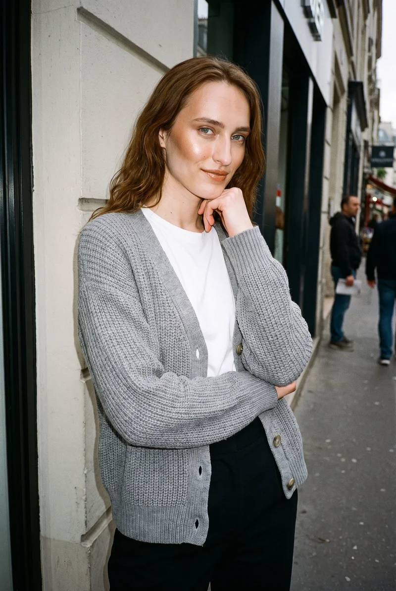 A 21-year-old Slavic woman wears a grey knit cardigan in an angled upper-body view, with one wrist raised near her jaw. She has a natural facial expression, and an urban street appears as the backdrop.