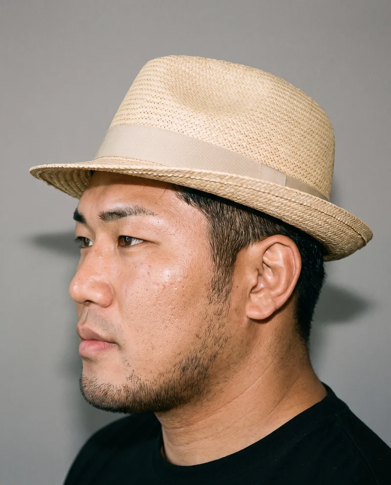 A 31-year-old Japanese man is shown from the head up in side view, wearing a beige straw fedora with his chin raised and a natural expression. A studio grey wall forms the backdrop.