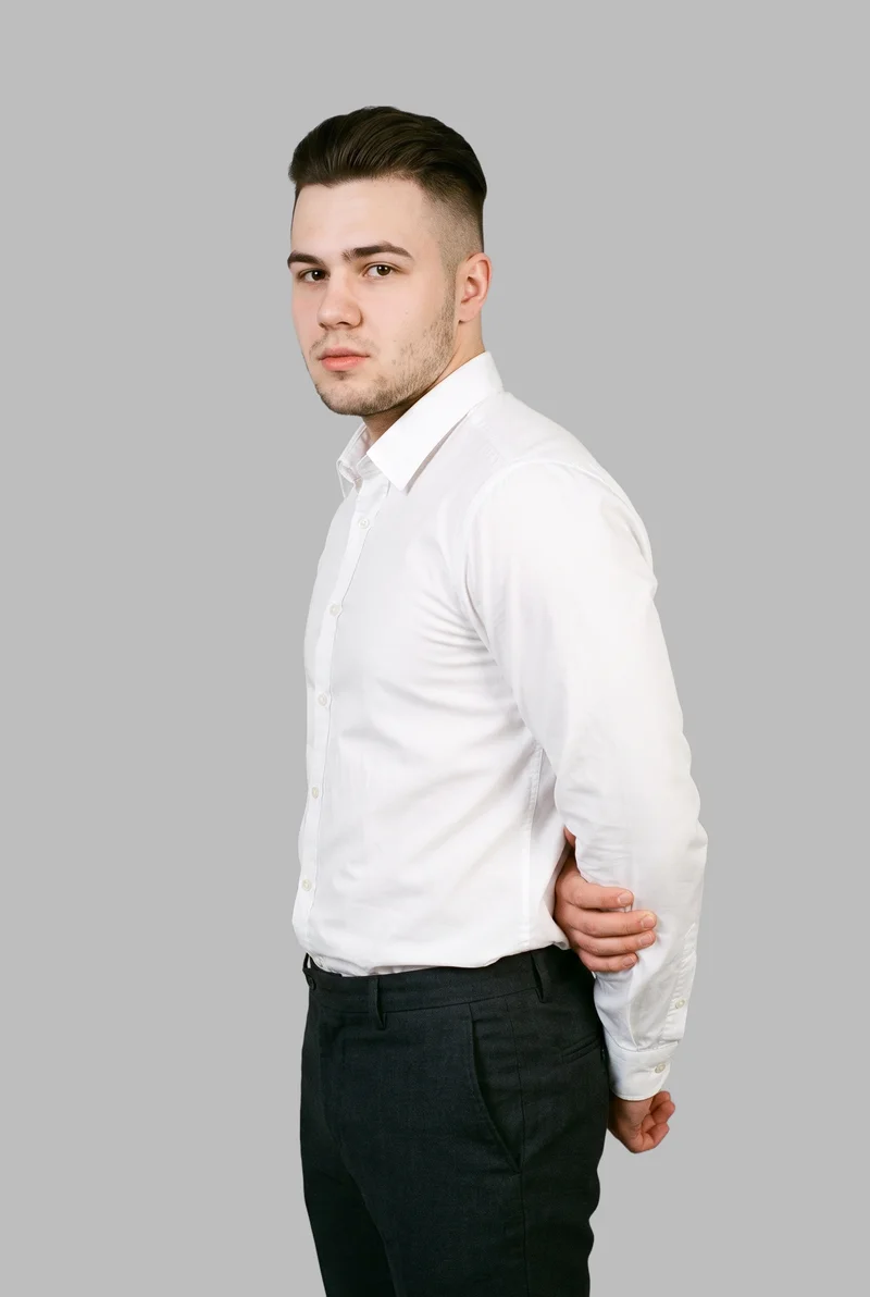 A 21-year-old Slavic male model wears a white button-down shirt against a dove-grey backdrop. He is shown in a three-quarter-length front three-quarter view with one elbow positioned behind his back and a neutral facial expression.