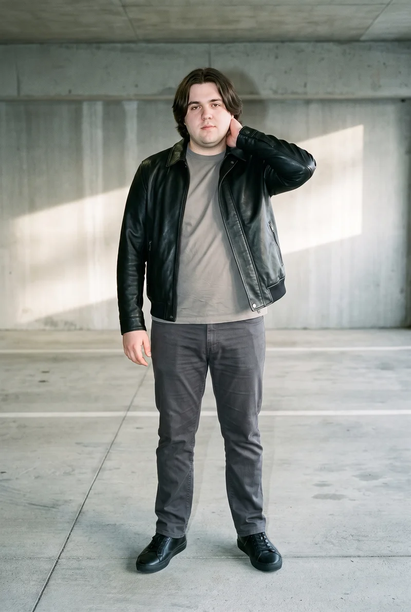 A 23-year-old Caucasian man wears a black leather bomber jacket in a full-body front view. He stands with one hand behind his neck and a neutral expression against a concrete parking garage backdrop.
