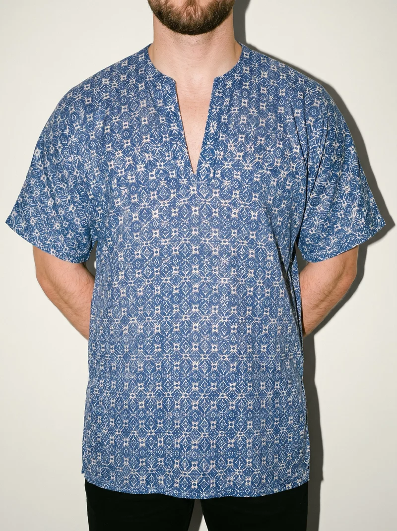 A male model wears a men's printed kaftan cover-up in a front-view torso composition, standing with his hands behind his back. An alabaster studio-wall backdrop fills the background.
