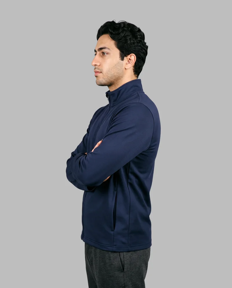 A 22-year-old Arab man wears a men's navy tracksuit top in a three-quarter-length side view. He poses in a self-hug with a confident facial expression against a dove grey flat-color backdrop.