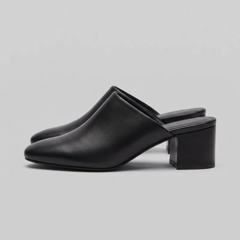 A pair of black closed-toe heeled mules is shown in a standalone, side-angled view. The shoes have covered insteps, open backs, rounded almond toes, padded footbeds with raised rims, thin outsole edges, and straight block heels. Their plain exterior has subtle fine-grain texture and stitched trim, with no visible straps, laces, buckles, logos, or embellishments.