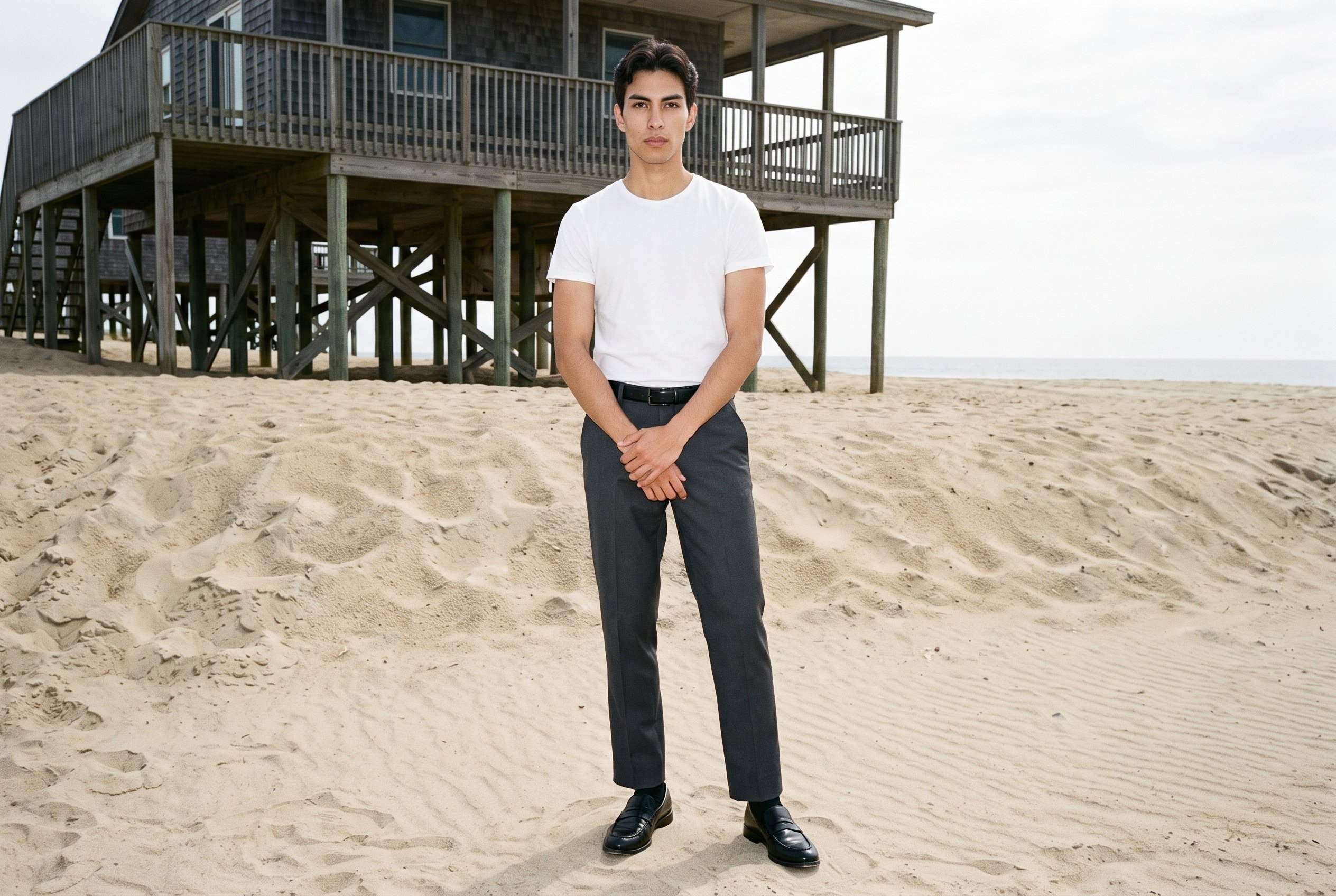A 20-year-old Hispanic male model wears charcoal tailored trousers in a full-body front view, standing with his hands in front and an editorial facial expression. An Outer Banks Beach House backdrop is visible behind him.