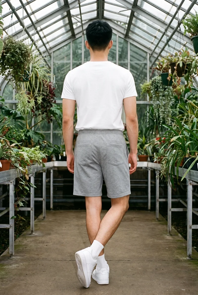 An 18-year-old Chinese male model is shown full body from the rear wearing grey gym shorts, with his ankles crossed. A greenhouse conservatory forms the backdrop.