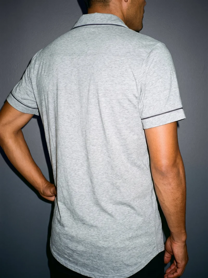 A grey nightshirt worn by a male model, shown in a torso crop from an angled rear view with one hand on his hip. A graphite studio-wall backdrop is visible.