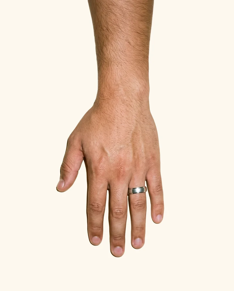 A man's hand and wrist are shown from above at rest, wearing a men's silver band ring. The subject is framed closely against a creamy white flat-color backdrop.