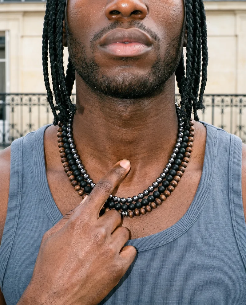 A man's neck shown from the front with a finger touching his throat, wearing a men's beaded layered necklace. A Paris balcony serves as the backdrop.