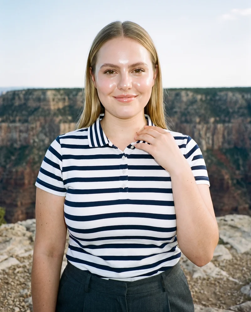 A 22-year-old Caucasian woman wears a women's striped cotton polo shirt in an upper-body front view. She is smiling with one hand at her collarbone, with a Grand Canyon rim backdrop.