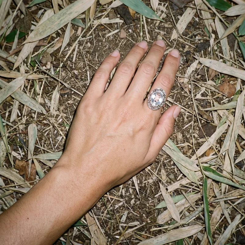 A silver cocktail ring is shown on a woman's hand and wrist from a top view, with the hand angled within the frame. A bamboo forest backdrop is visible behind the jewelry.