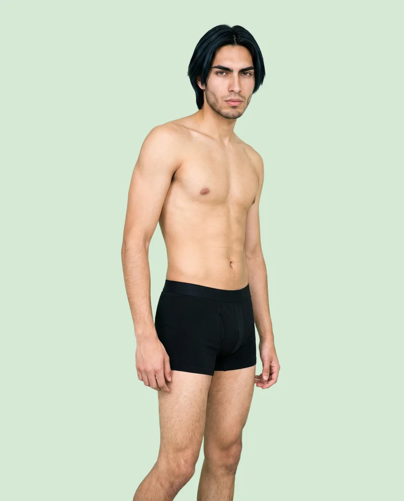 An 18-year-old Hispanic male model stands in a relaxed pose, shown full body from a front three-quarter angle, wearing black boxers. The expression is natural, with a flat pistachio backdrop.