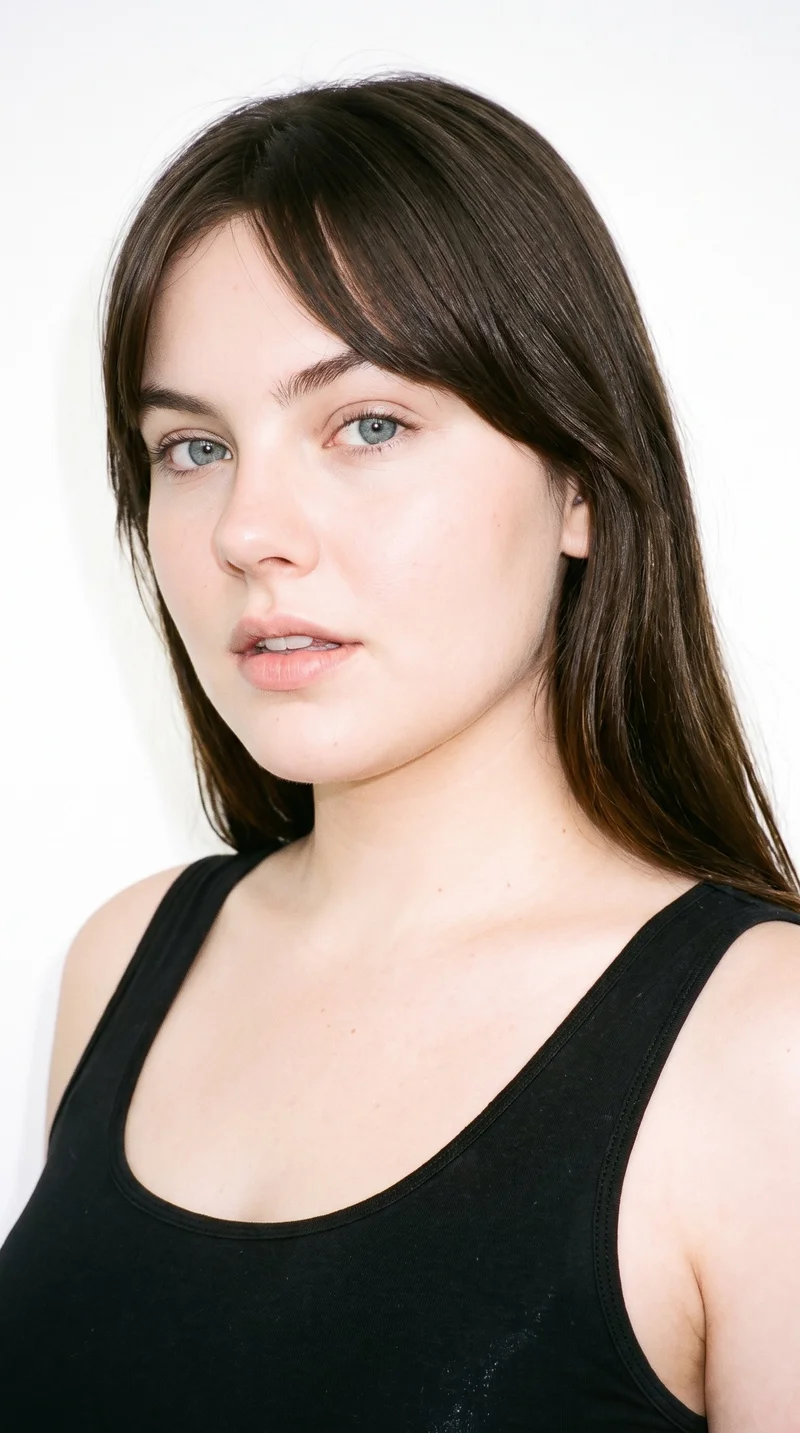 This 24-year-old Caucasian woman has lightly tanned fair skin with a neutral undertone. Her face has soft, youthful features, full cheeks, and rounded contours. She has medium-thickness eyebrows, blue eyes with a cool undertone, and thin lips. Her dark-brown hair is styled with longer centre-parted bangs that frame both sides of her face. No facial hair is specified.
