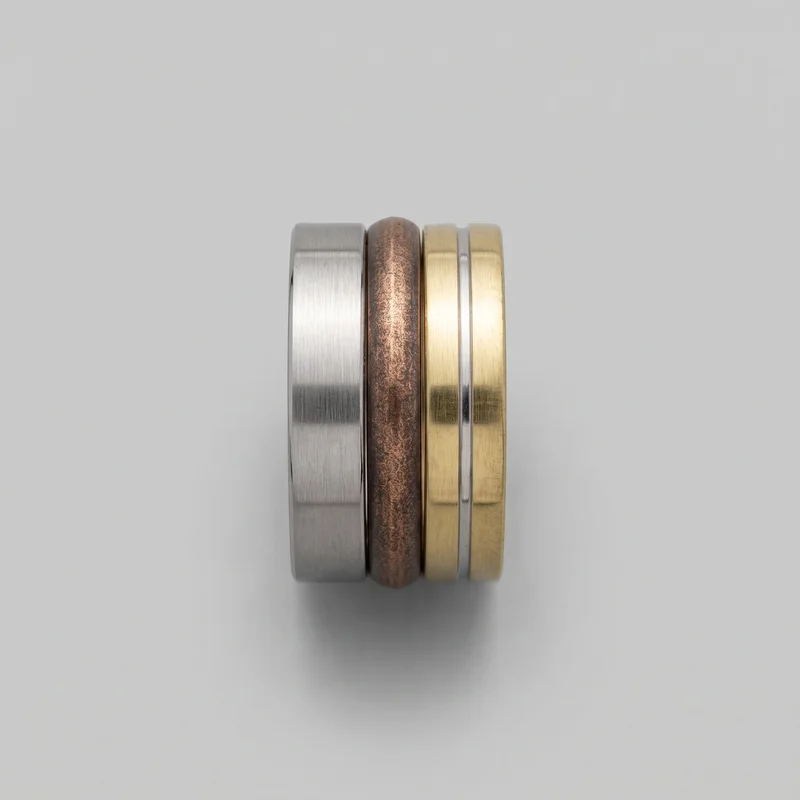 Multiple mixed-metal finger bands are stacked upright side by side in a close standalone product view. The set includes a flat-faced brushed band with softly rounded edges, a rounded domed band with a mottled hammered-looking surface, and a flat-faced band with a recessed contrasting center channel. No stones, logos, text, or figurative embellishments are visible.