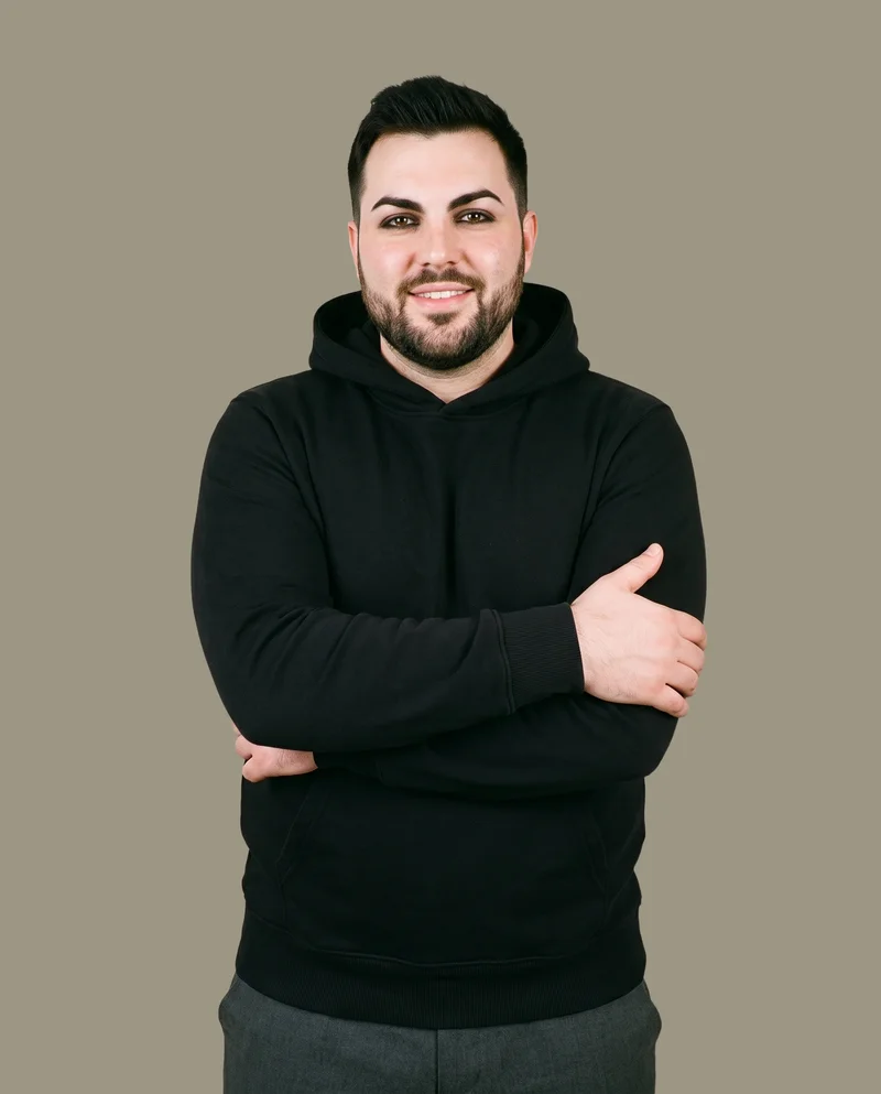 A 28-year-old Caucasian man is shown from the front in an upper-body view wearing a black pullover hoodie. He is smiling and holding his elbow, with a warm grey flat-color backdrop.