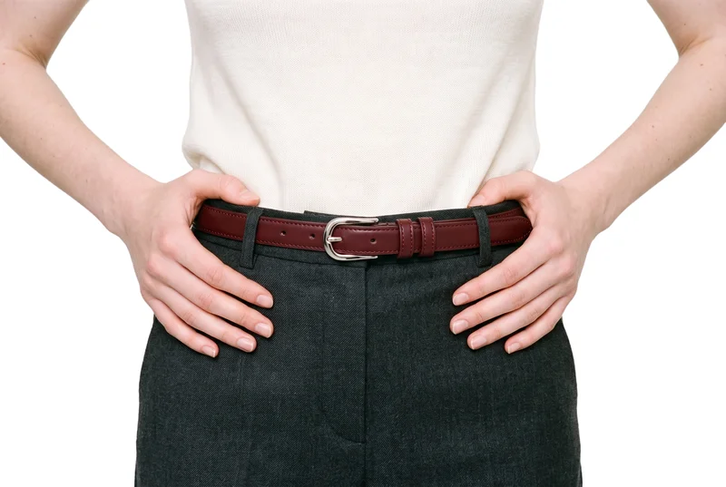 A woman wears an oxblood leather dress belt around her waist, shown in a front view with her hands resting on her waist. The waistline is framed against a pure white backdrop.