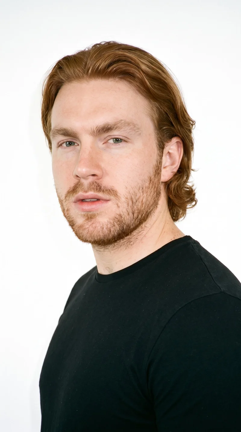 A 25-year-old Caucasian man with very pale skin and a recorded warm-golden undertone has a softly structured, refined face with even features. His eyebrows are thin, and his eyes are blue with a cool undertone. He has full lips and dense, heavy stubble forming a dark, defined layer across the beard area. His vivid red hair is medium to long, brushed back with body and movement extending past the ears.