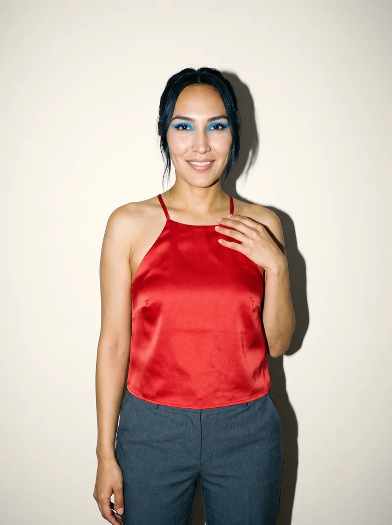 A 25-year-old Indigenous American woman is shown from the upper body in a front view, wearing a red satin halter top. Her fingers are held at her chest, and she is smiling against a creamy white studio-wall backdrop.