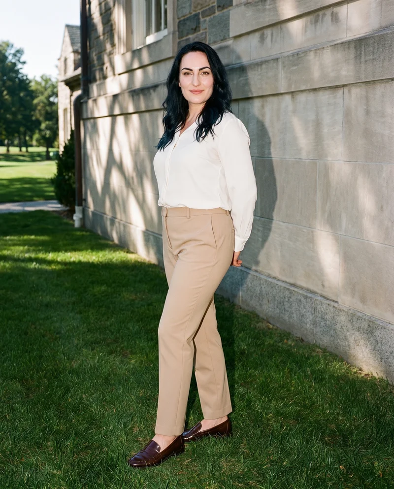 A 37-year-old Slavic woman wears beige suit trousers, shown full body from a front three-quarter angle with her hands behind her back and a natural facial expression. A country club backdrop is visible.