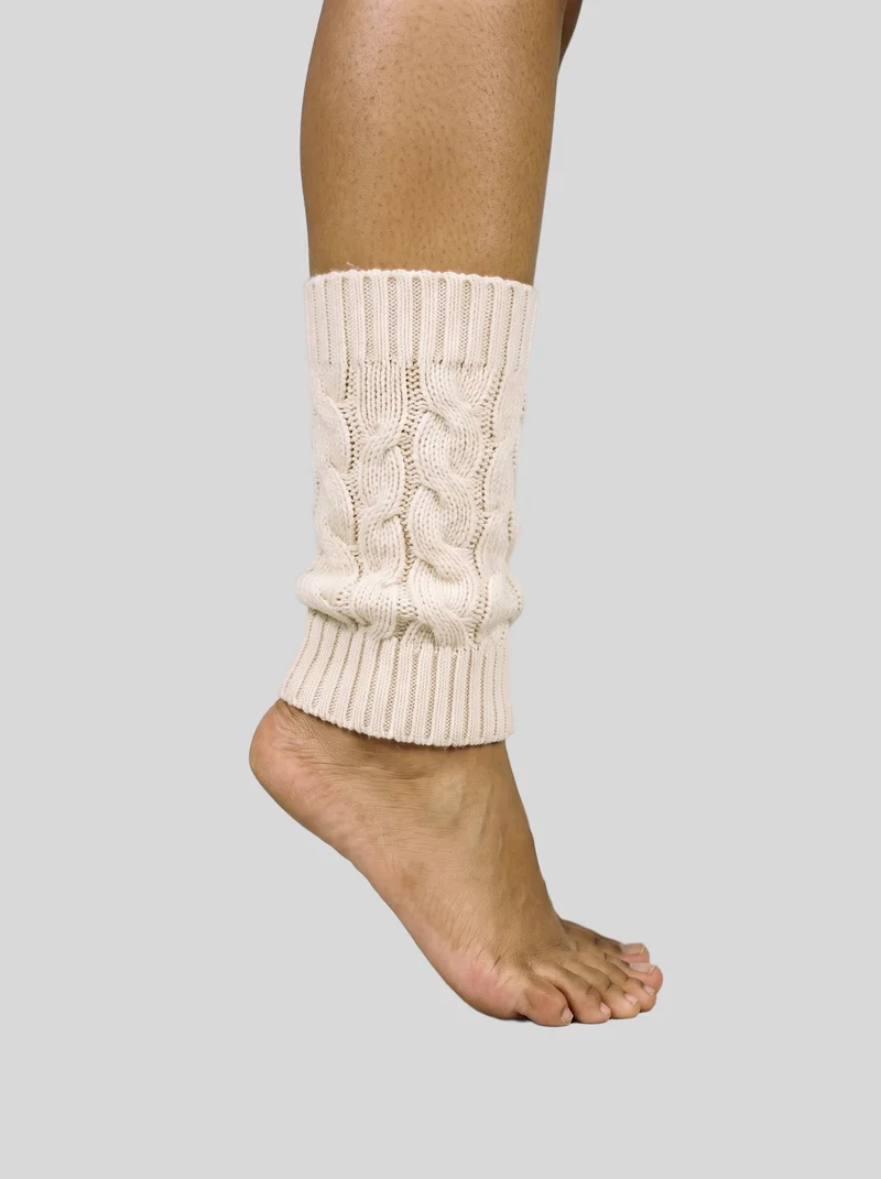 Cream cable-knit leg warmers worn on the lower legs, shown in an ankle-level side view while the wearer stands on tiptoe. The image uses a flat pearl grey backdrop.