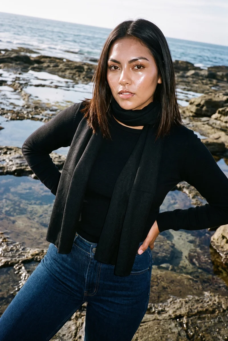 A 23-year-old Hispanic woman is shown from the upper body at an angled front view, wearing a black pashmina shawl with her hands on her hips and a neutral expression. A rocky beach serves as the backdrop.