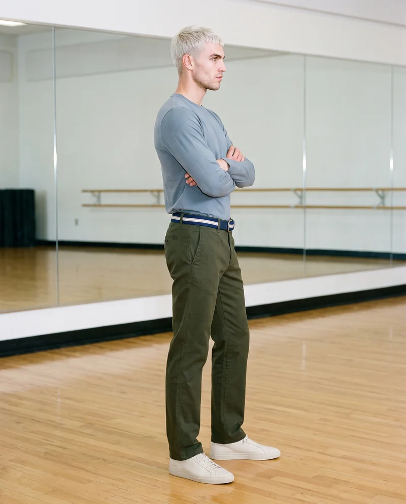 A 29-year-old Caucasian man is shown full body in side view wearing a striped woven belt. He stands in a self-hug pose with a confident facial expression against a dance studio backdrop.