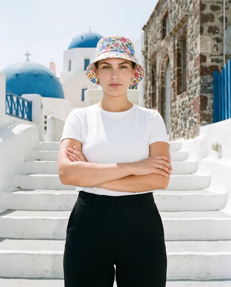 A 29-year-old Caucasian woman wears a women's floral print bucket hat in an upper-body, front-facing view. Her arms are crossed, and the image uses an editorial facial expression against a Santorini white steps backdrop.