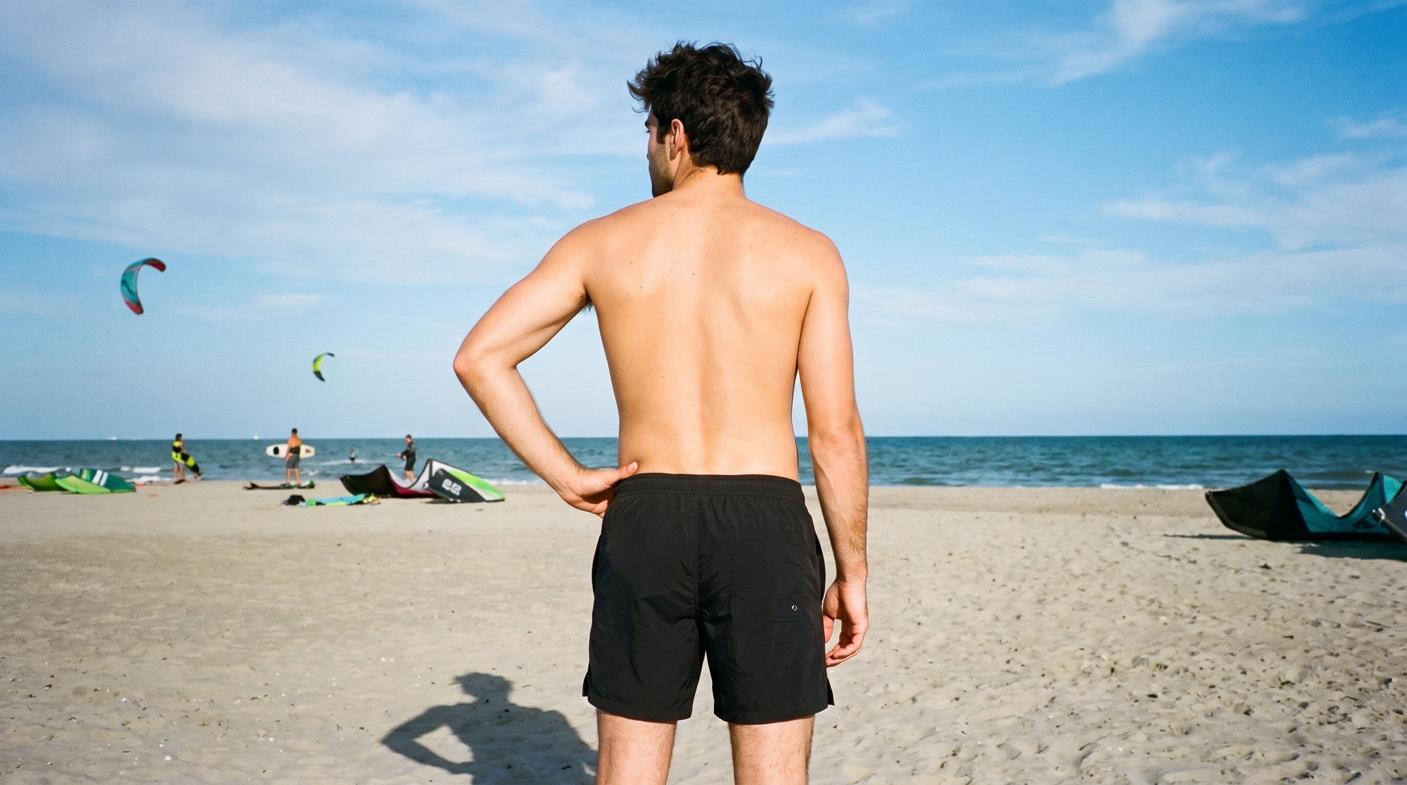 A 24-year-old Caucasian man wears black swim trunks in a three-quarter-length rear view, with one hand on his hip. A kitesurfing beach backdrop is visible behind him.