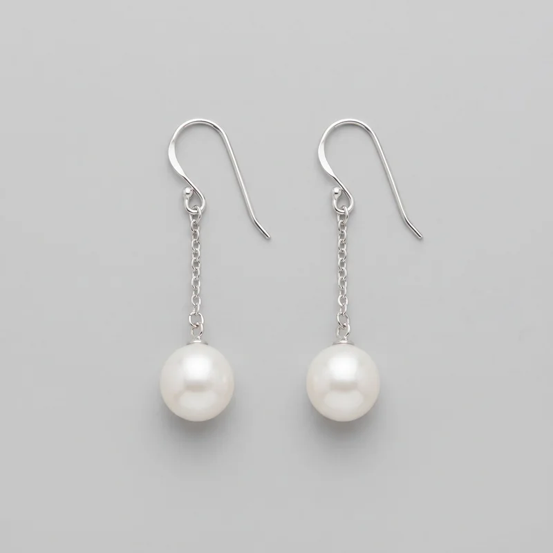 A matched pair of pearl drop earrings is laid flat in a standalone product view. Each earring has a slim curved fish-hook ear wire, a small loop connector, a short linked chain drop, and a smooth spherical bead pendant, with the lower edges of the pendants close to the frame.