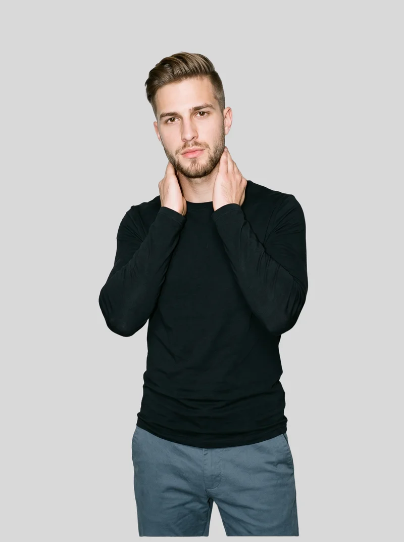 A 22-year-old Caucasian male model wears a men's black compression base layer in an upper-body view from a front three-quarter angle. His hands are positioned on his neck, and he has a neutral facial expression. A pearl grey flat-color backdrop frames the studio cutout.