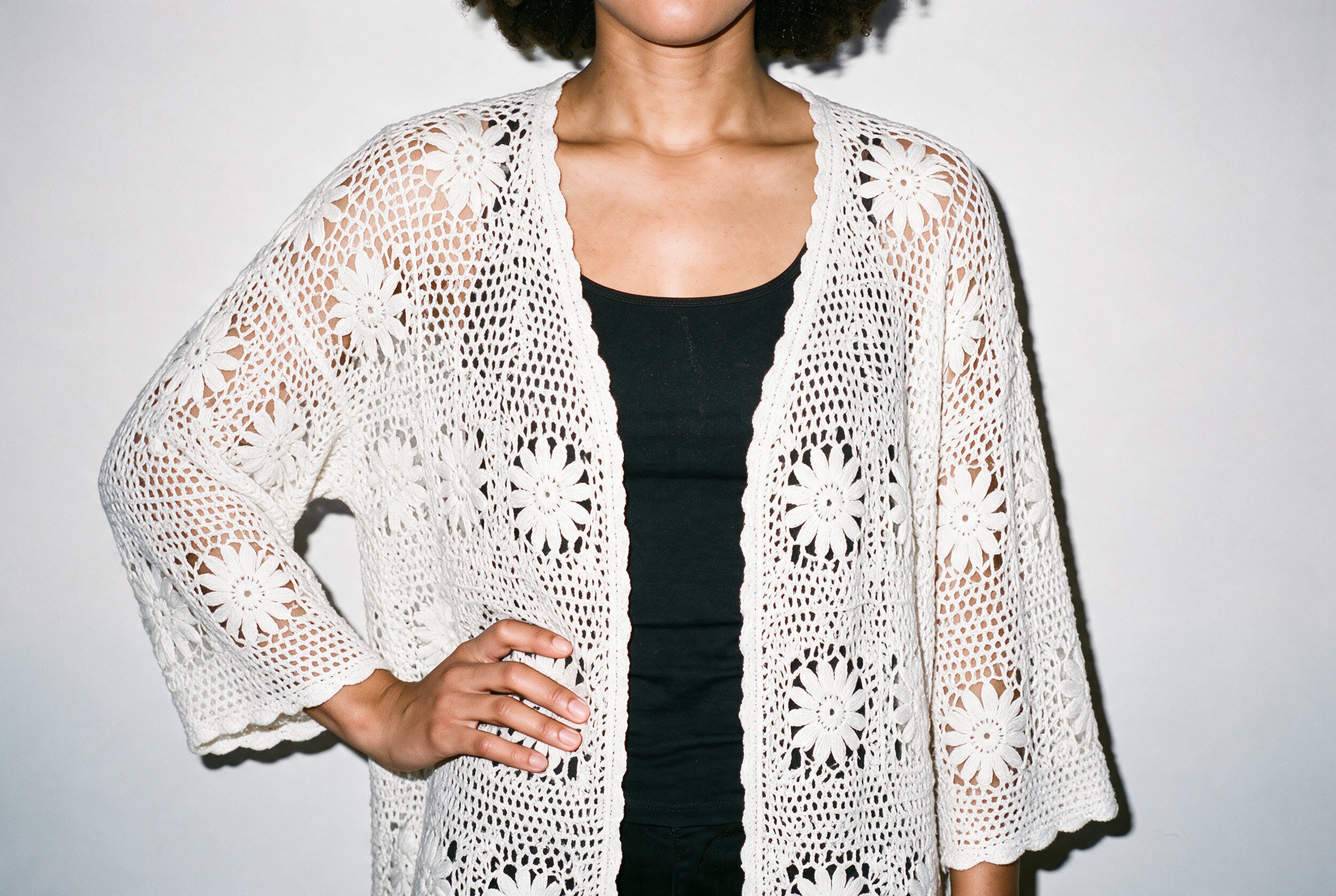 A woman wearing a white crochet cover-up is shown from the torso in a front view, with one hand on her hip. A fog grey studio-wall backdrop frames the garment.